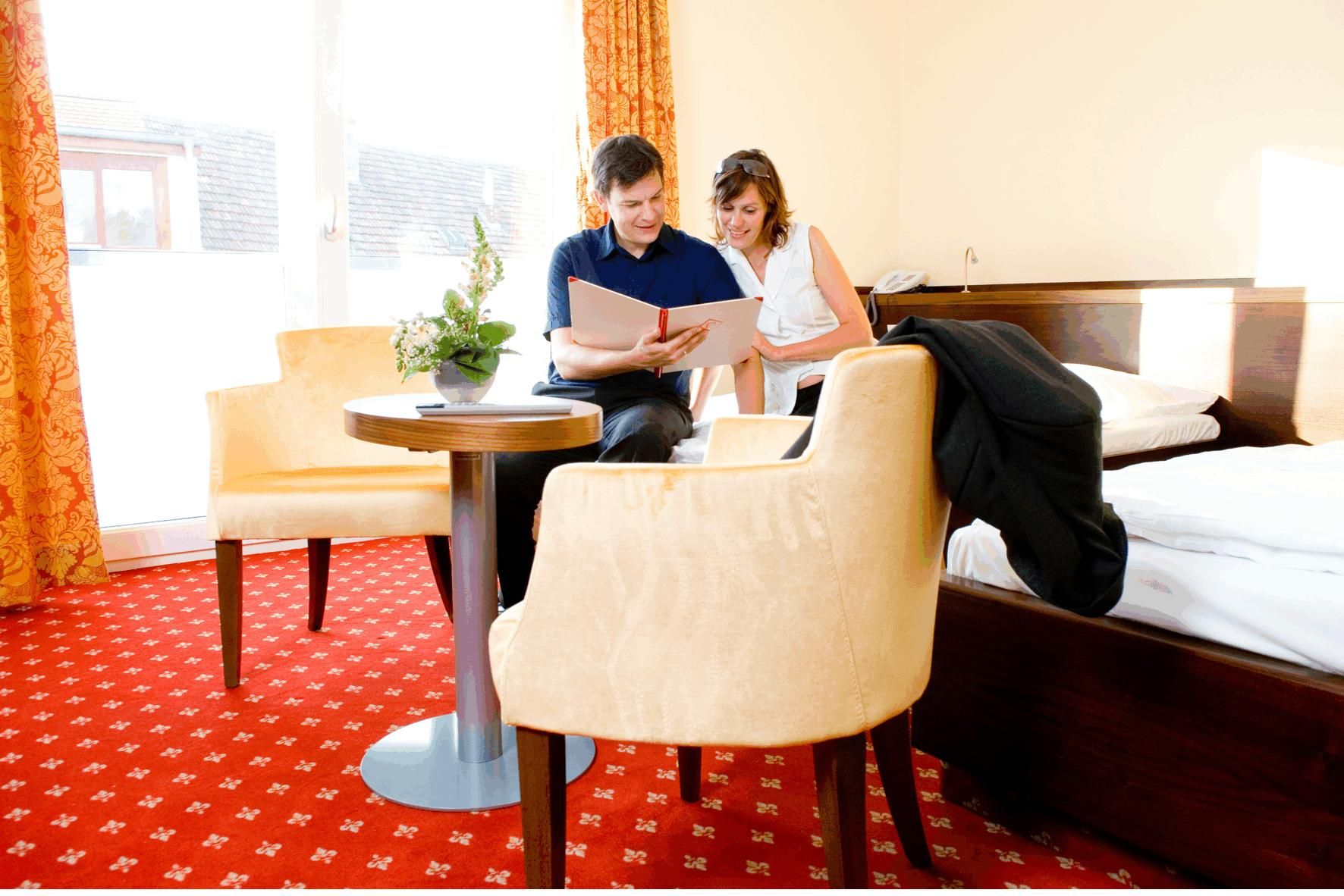 A couple is sitting on a bed in a hotel room, looking into a book. The room has red carpets and yellow curtains.