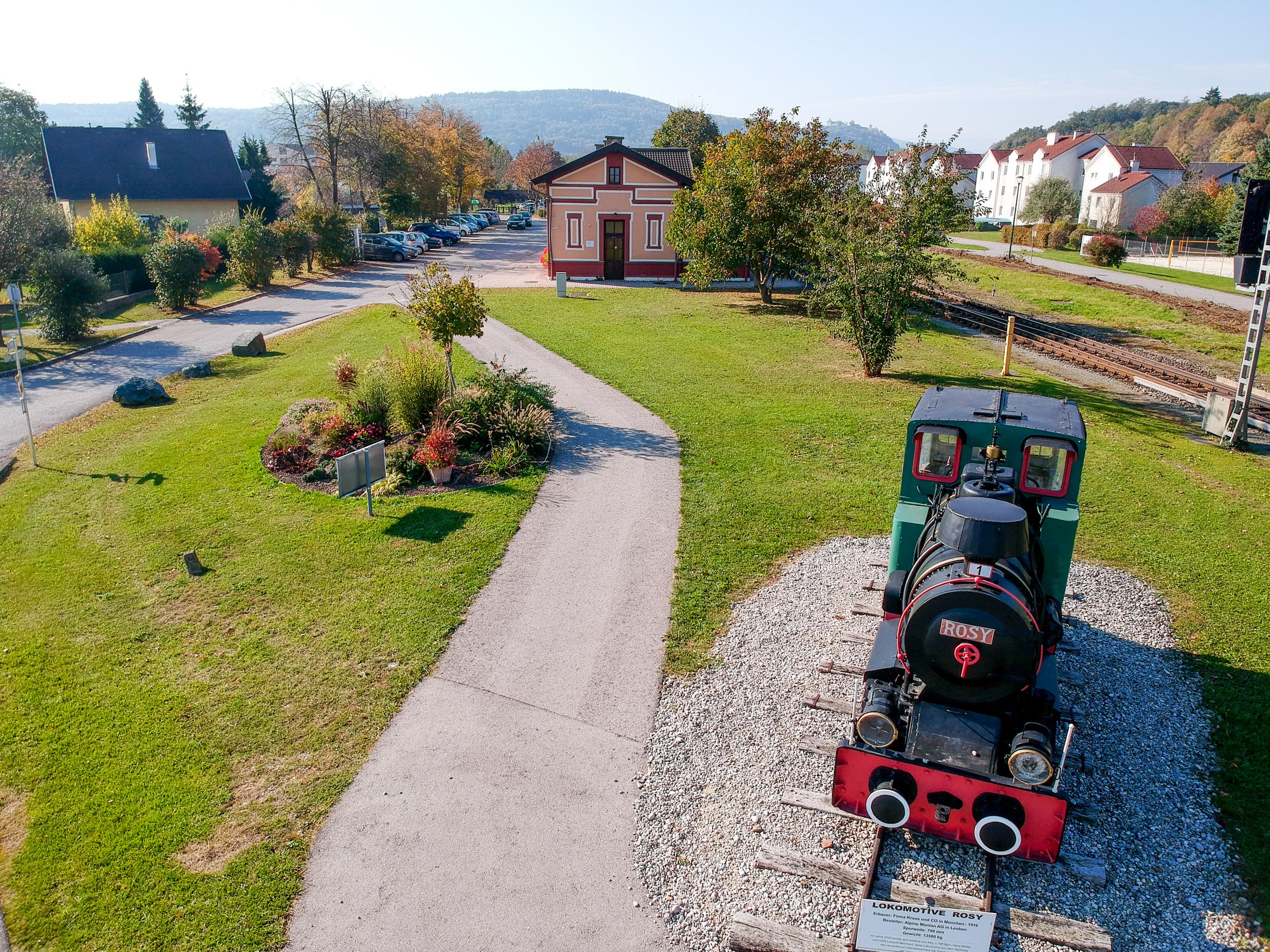 A small historic locomotive called 'Rosy' stands on a short section of track in a green park next to a small station building.