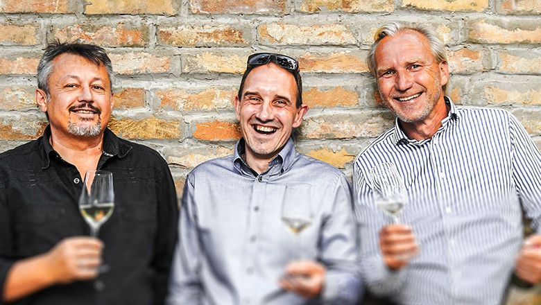 Three men stand in front of a brick wall holding glasses of wine.
