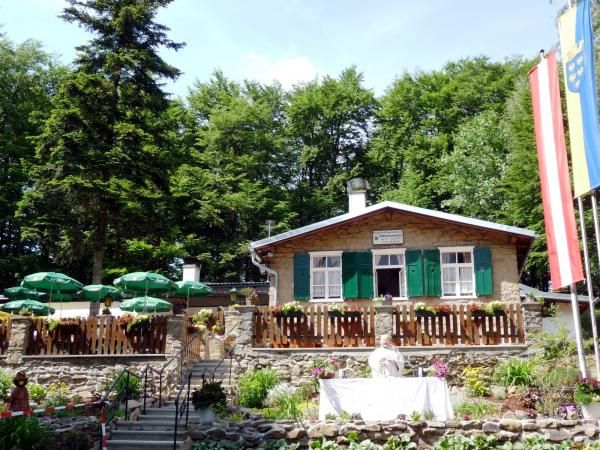 A mountain hut with green shutters, surrounded by trees, with a garden and parasols in the foreground.