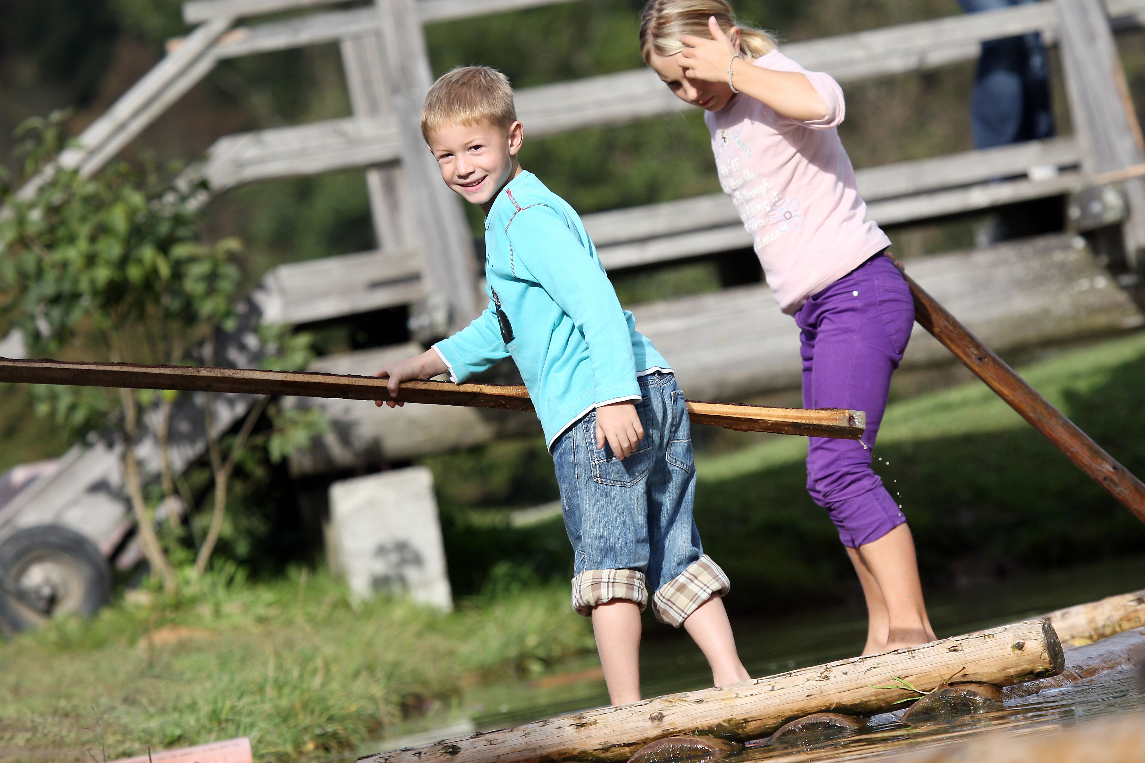 Two children balance on a wooden raft over water, in the background a wooden bridge.