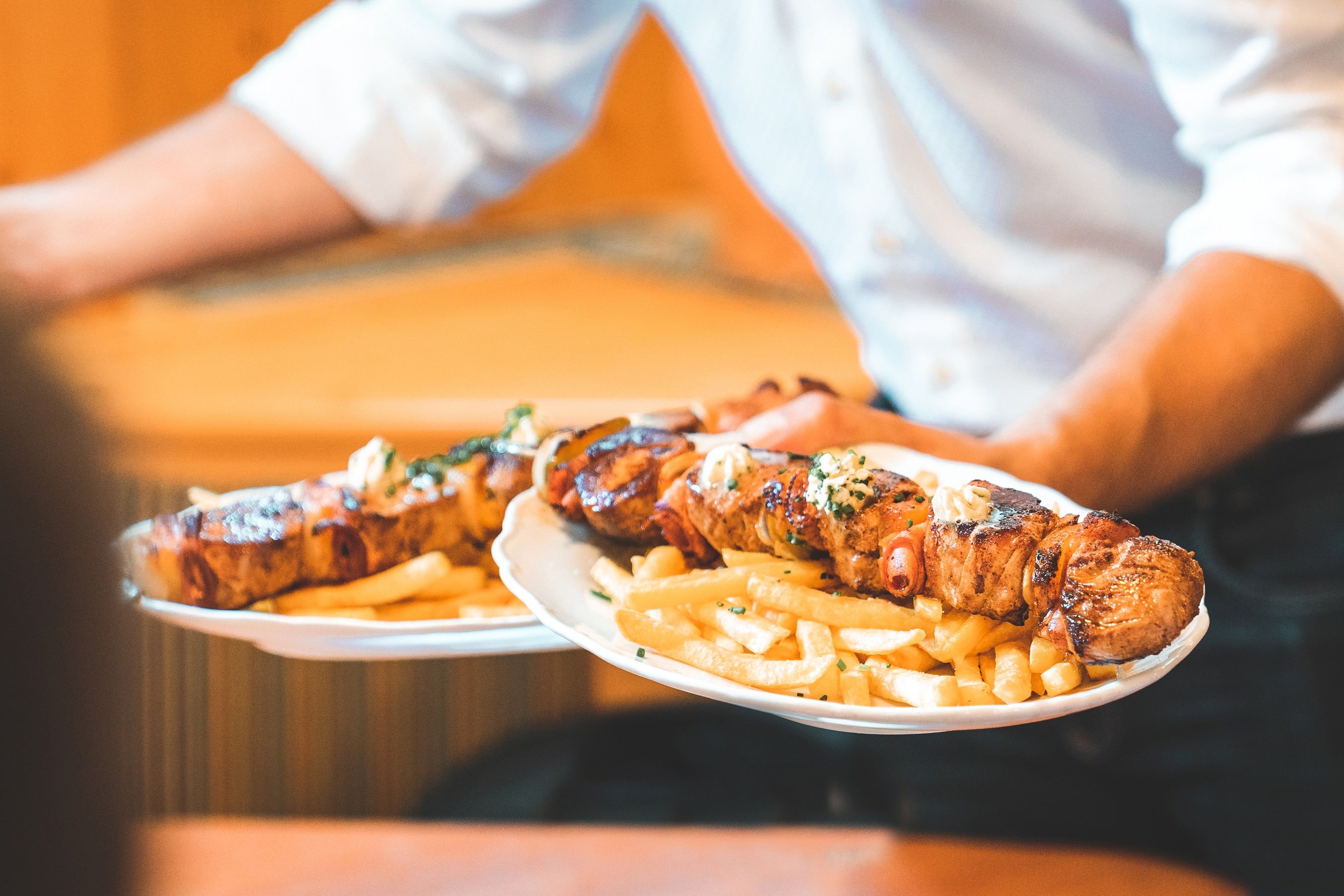 A waiter serves two plates of grilled meat skewers and chips.