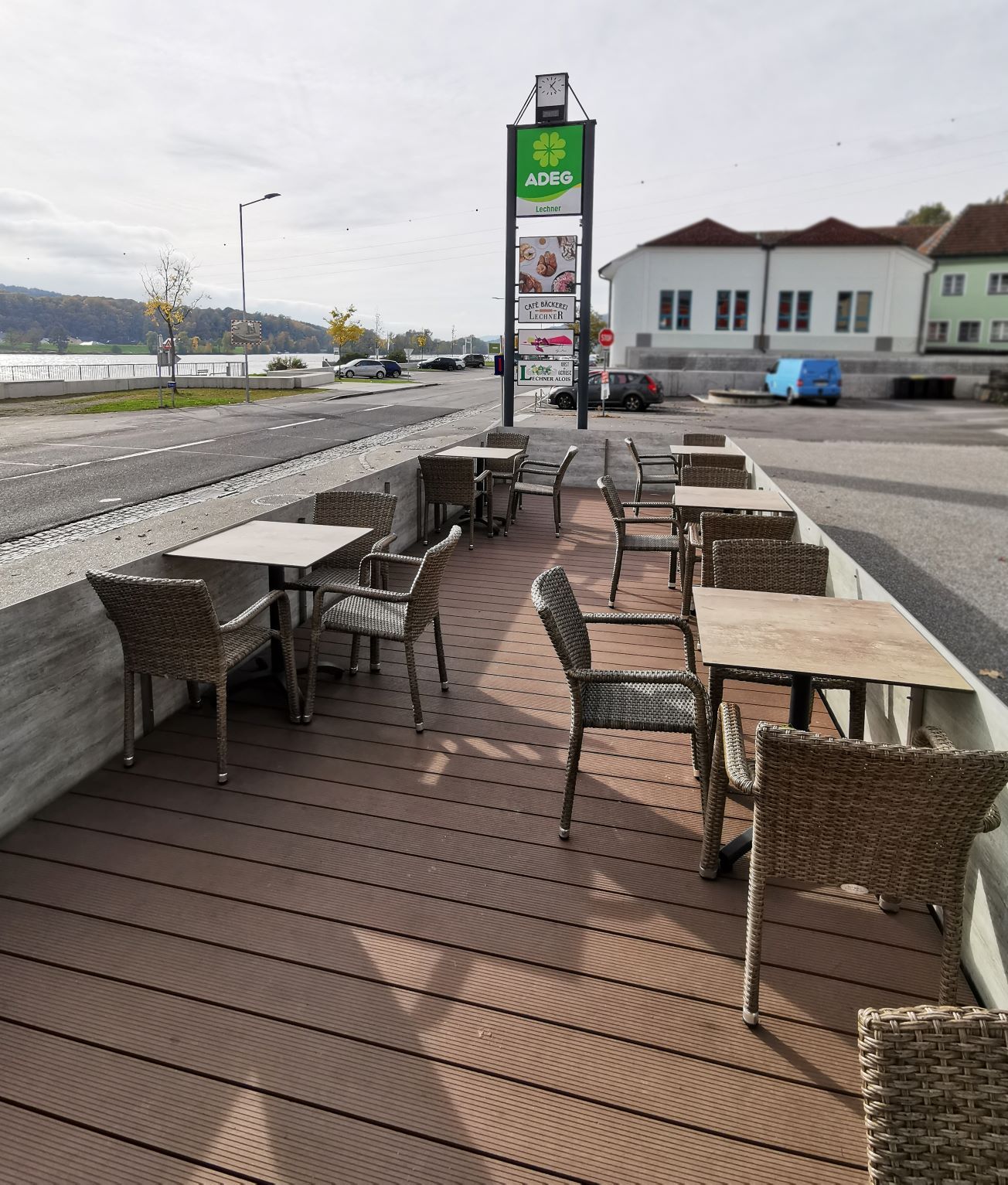 Outside area of a café with empty tables and chairs, next to a street and a sign saying 'ADEG'.