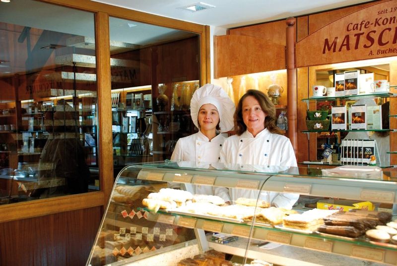 Two women in pastry chef uniforms stand behind a counter with pastries in a bakery.