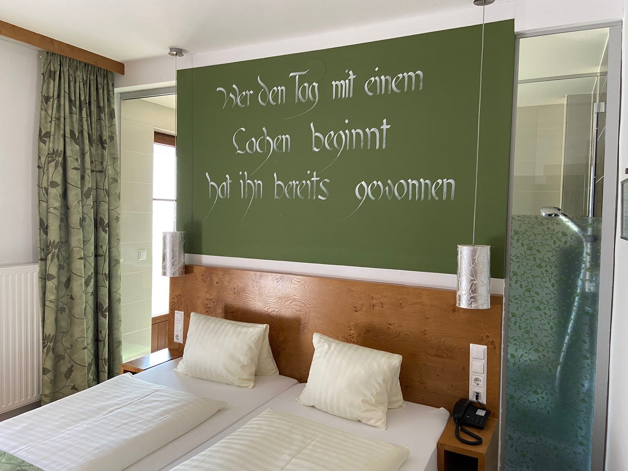 A double room with two beds, a green wall with a slogan, curtains and a shower with a glass wall.