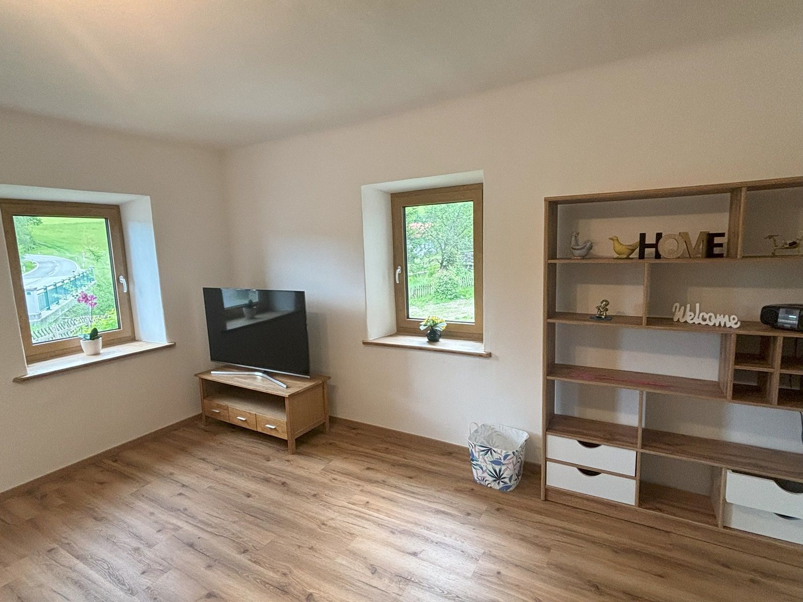 A living room with a wooden floor, a TV on a table, two windows and a shelf with decorations.