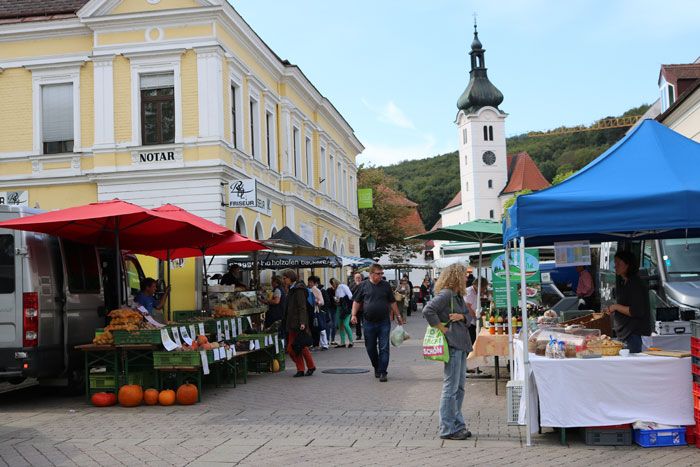 A busy farmers' market with stalls and visitors in front of a yellow building and a church in the background.