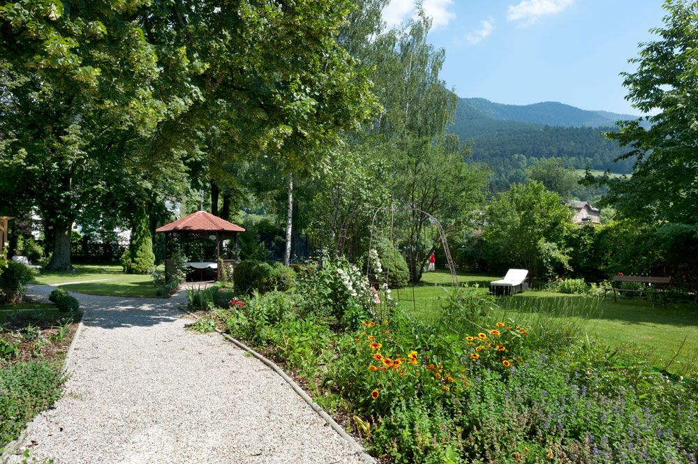 A well-kept garden with a gravel path, flower beds, a pavilion and sun loungers against a mountain backdrop.
