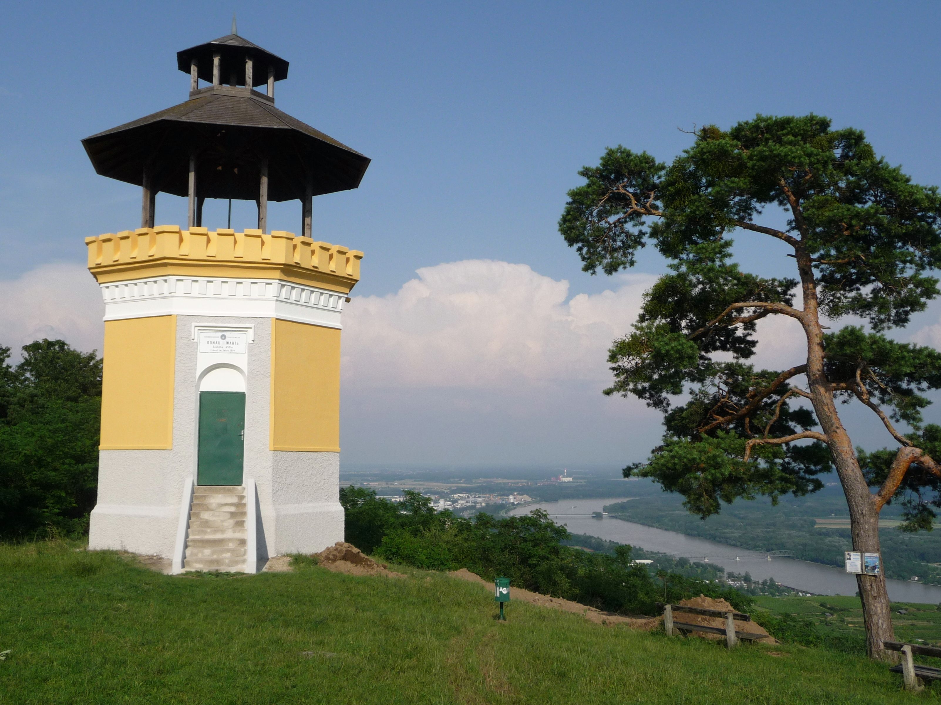 A yellow observation tower with a dark roof stands on a hill with a view of the Danube river and a town in the background.
