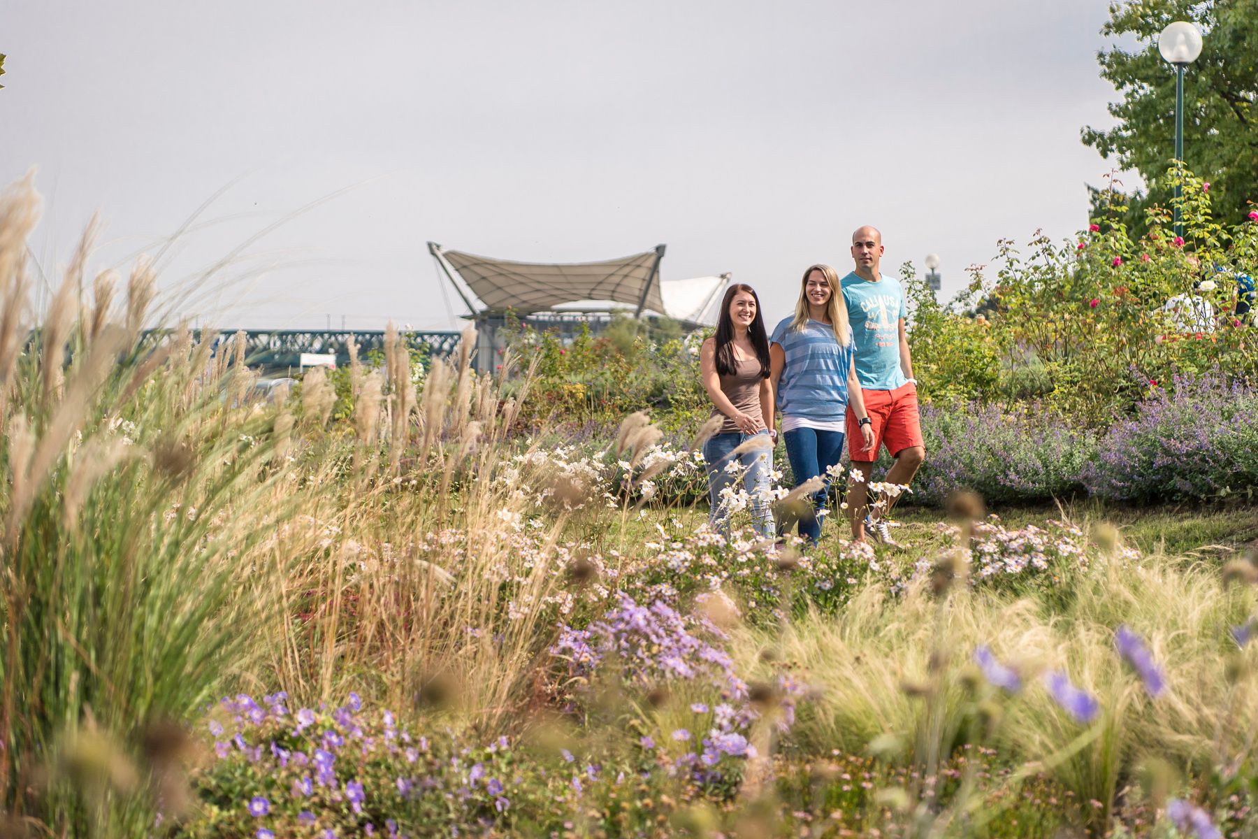 Three people stroll through a blooming garden with grasses and flowers, a bridge and a modern roof in the background.