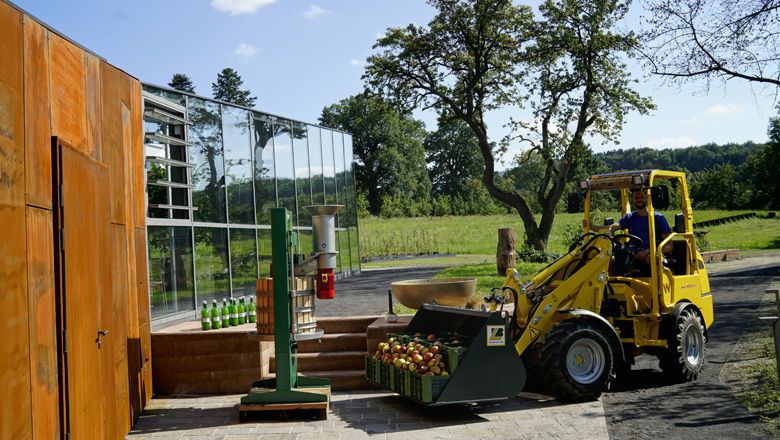A yellow electric Hoftrac transports crates of apples in front of a modern building on a farm.