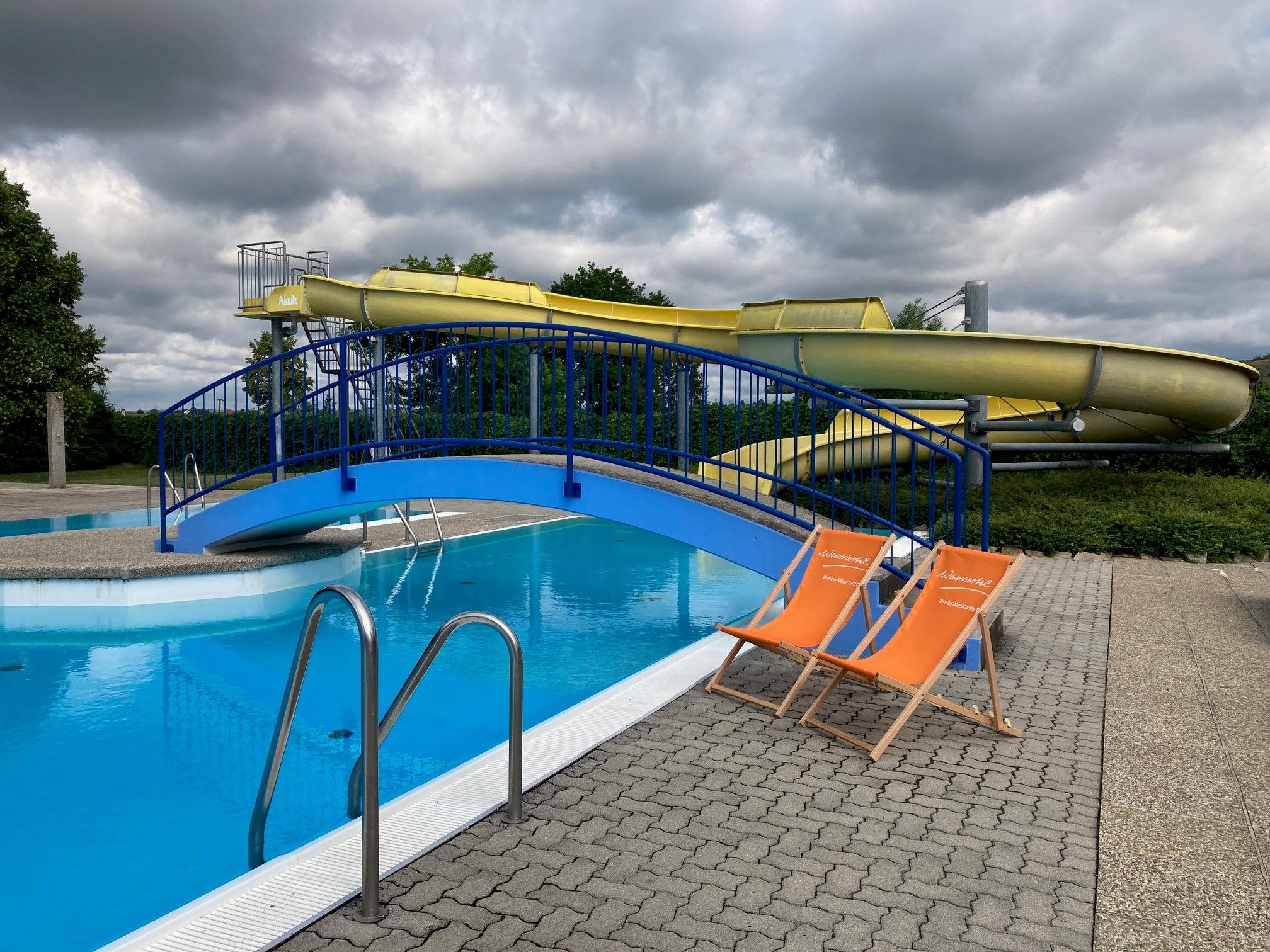 Two orange deckchairs stand next to a swimming pool with a blue bridge and a yellow water slide in the background.