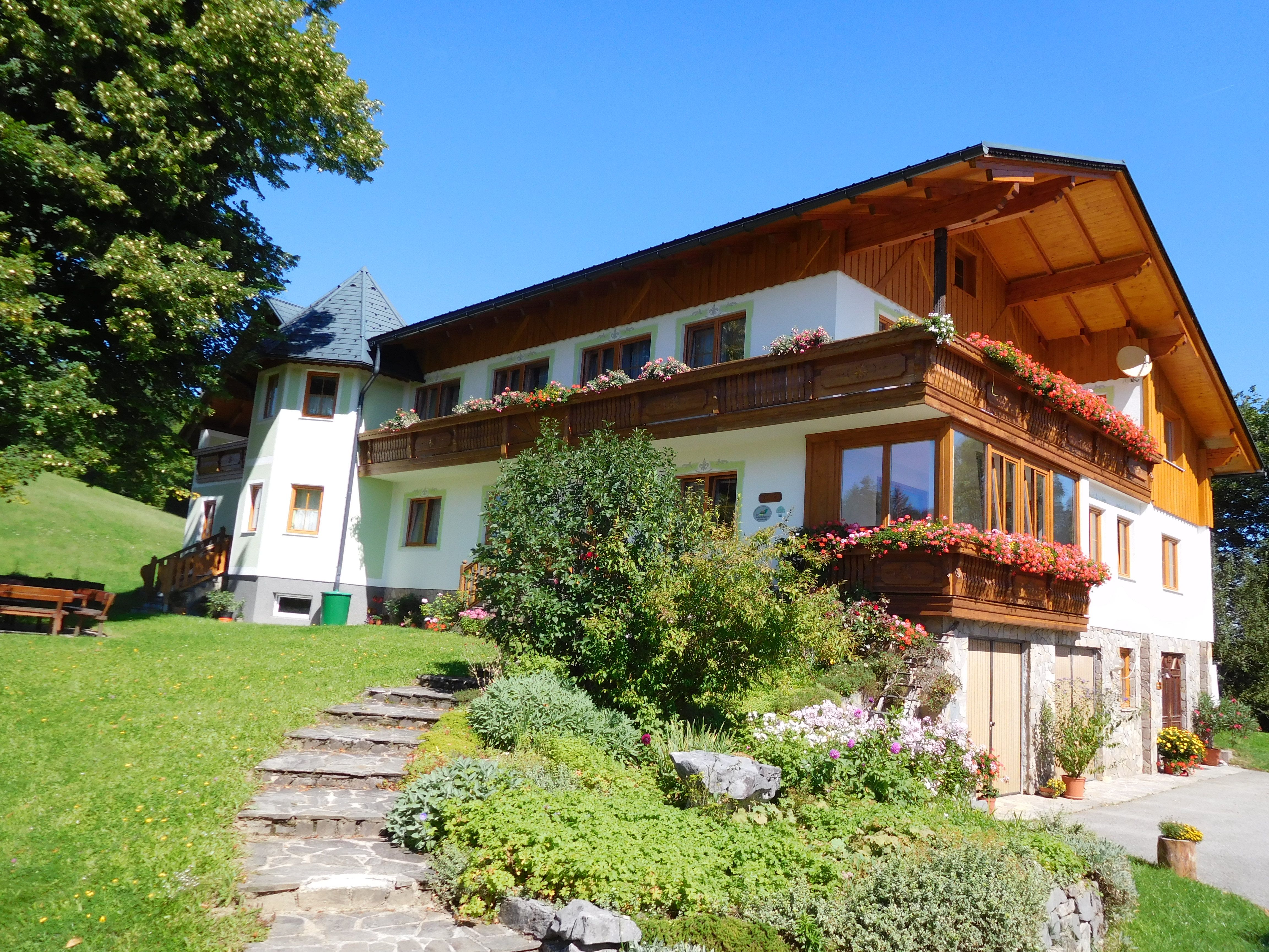 A traditional farmhouse with a wooden balcony and blooming flowers, surrounded by green countryside and blue skies.