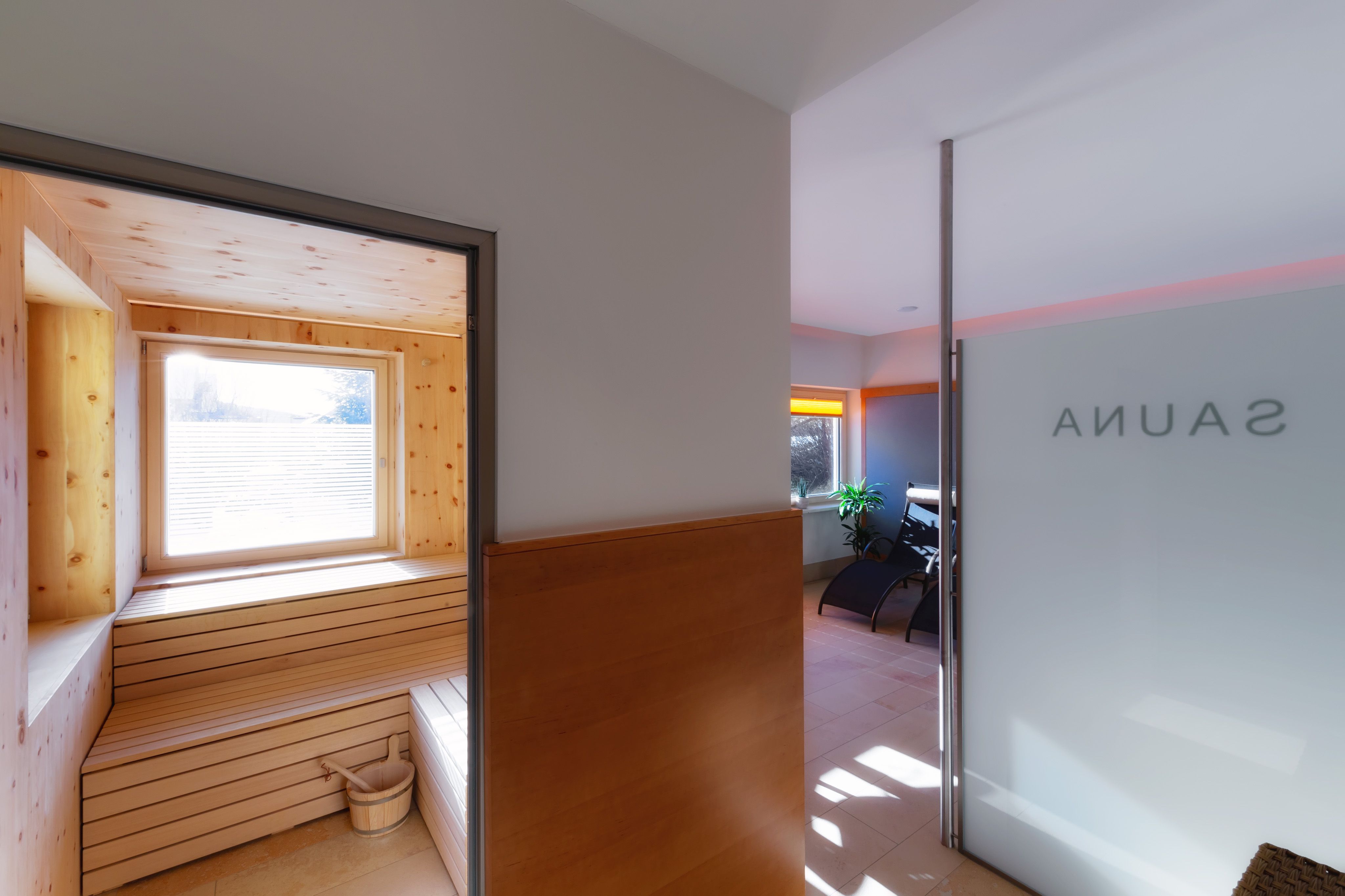 Interior view of a sauna with wooden paneling and window, next to it a relaxation room with loungers.