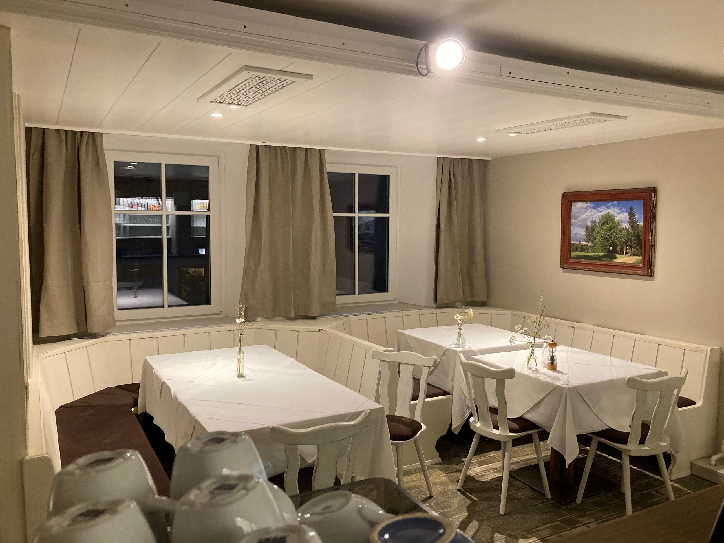 A cozy guest room with two set tables, white tablecloths and chairs. A landscape painting hangs on the wall and curtains can be seen through the windows.