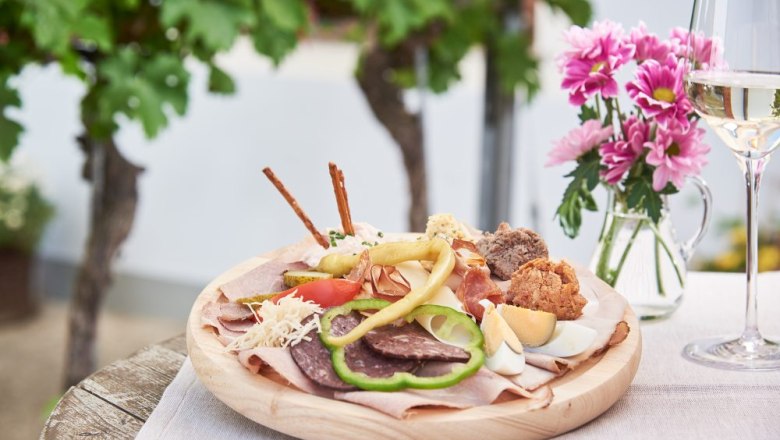 A plate with various wine tavern specialties, a glass of white wine next to it and pink flowers on an outdoor table.