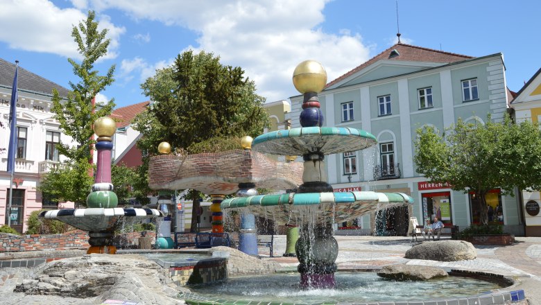 A colorful Hundertwasser-style fountain with golden balls and flowing water, surrounded by buildings and trees.