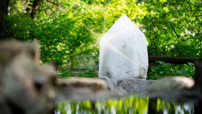 A large white crystal stands in a green forest, surrounded by plants and reflected in a small pond.