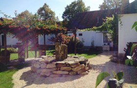 A sunny garden with a stone circle and a small fountain in the middle, surrounded by white buildings and green plants.