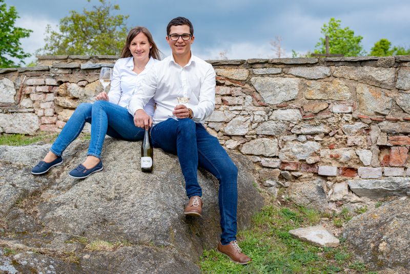 A man and a woman are sitting on a rock in front of a stone wall, holding wine glasses and a bottle of wine.
