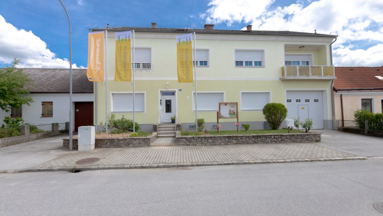 Exterior view of a two-story building with a yellow facade and two flags in front of it.