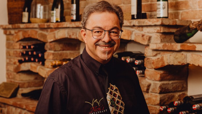 A man with glasses and a moustache stands smiling in front of a wine rack in a restaurant.