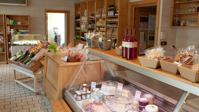 Interior view of a delicatessen with wooden shelves, vegetables, bottles of wine and a refrigerated counter with cheese and spreads.