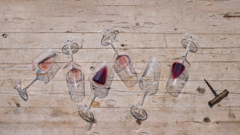 Six overturned wine glasses with red wine on a wooden table, a corkscrew next to them.