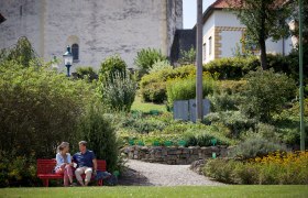 A couple sits on a red bench in a garden in front of the fortified church in Bad Sch&ouml;nau.
