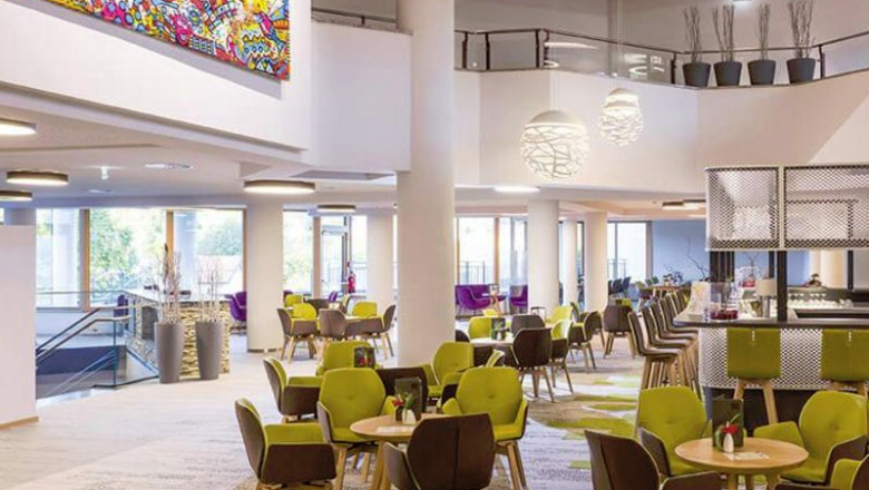 Modern hotel lobby with carpeted floor, round tables, green chairs, large lamps and a mural.