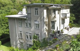 Large, multi-storey house with gray facade, surrounded by trees and garden.