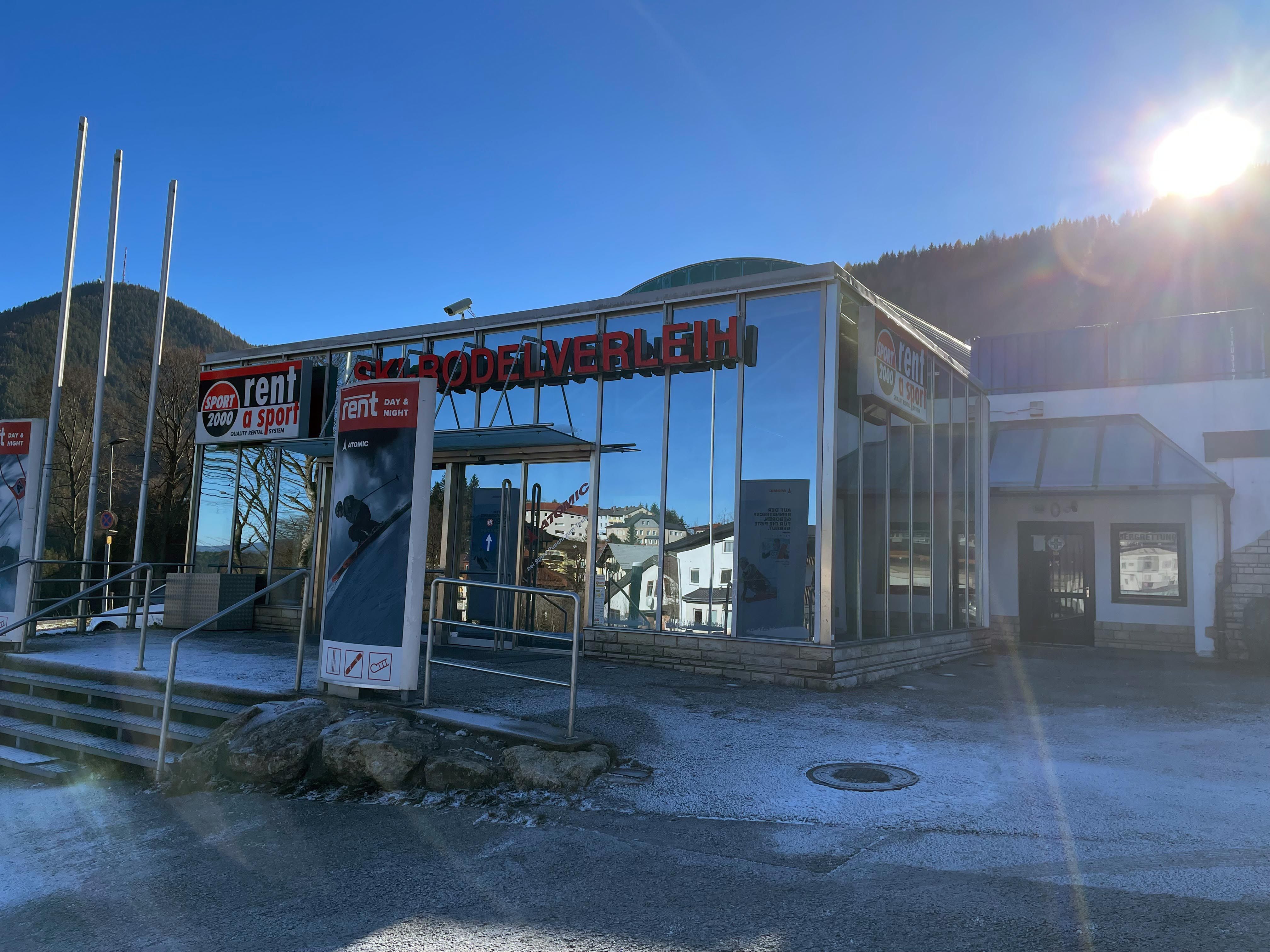 Exterior view of a ski rental shop with glass façade and advertising signs, surrounded by mountains and bright sunshine.