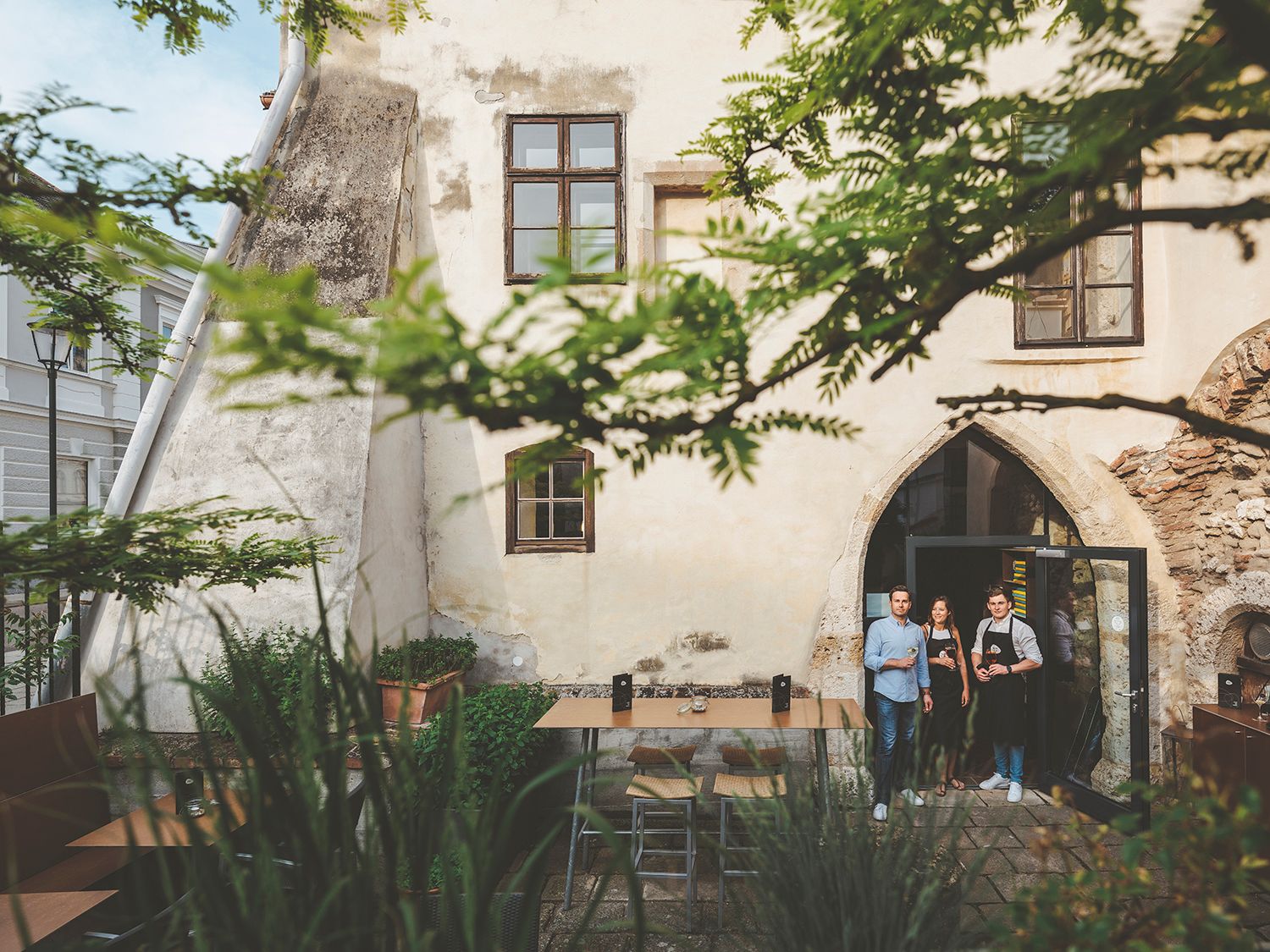 Three people are standing in a historic courtyard in front of a building with a Gothic arched window.