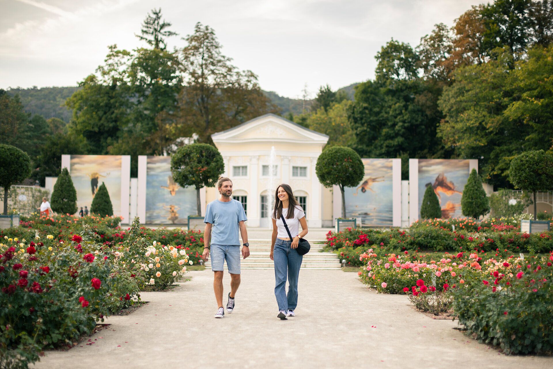 Zwischen blühenden Rosen im Doblhoffpark Baden spazieren eine Frau und ein Mann, vertieft in ein Gespräch. Im Hintergrund erhebt sich die eindrucksvolle Fotoausstellung von La Gacilly Baden Photo Baden, deren große Bilder das Szenario bereichern.