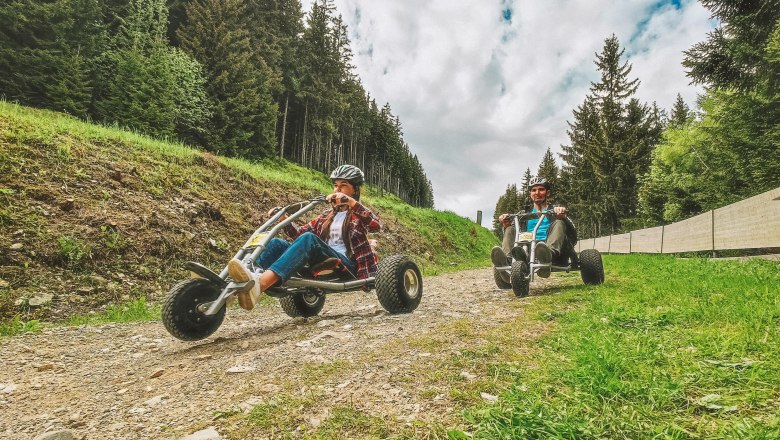 Two people are riding mountain carts on a gravel path in the forest.