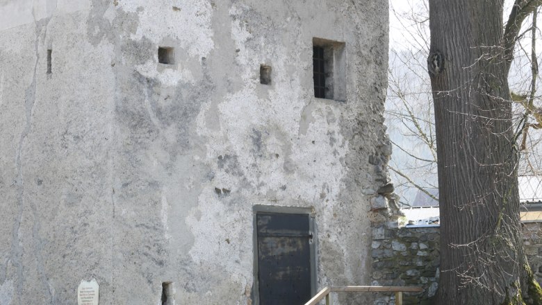 An old tower with a moss-covered roof and a tree next to it. Three people are sitting at a table while two people walk past.
