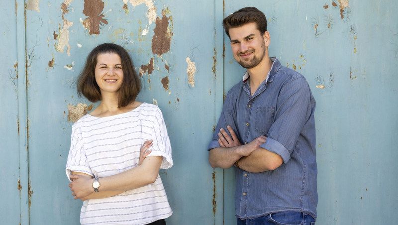 Two people are standing in front of a blue, worn wall.