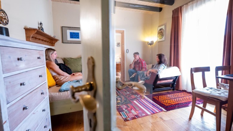 Three people sit in a cozy living room with a wooden floor and colorful carpet.