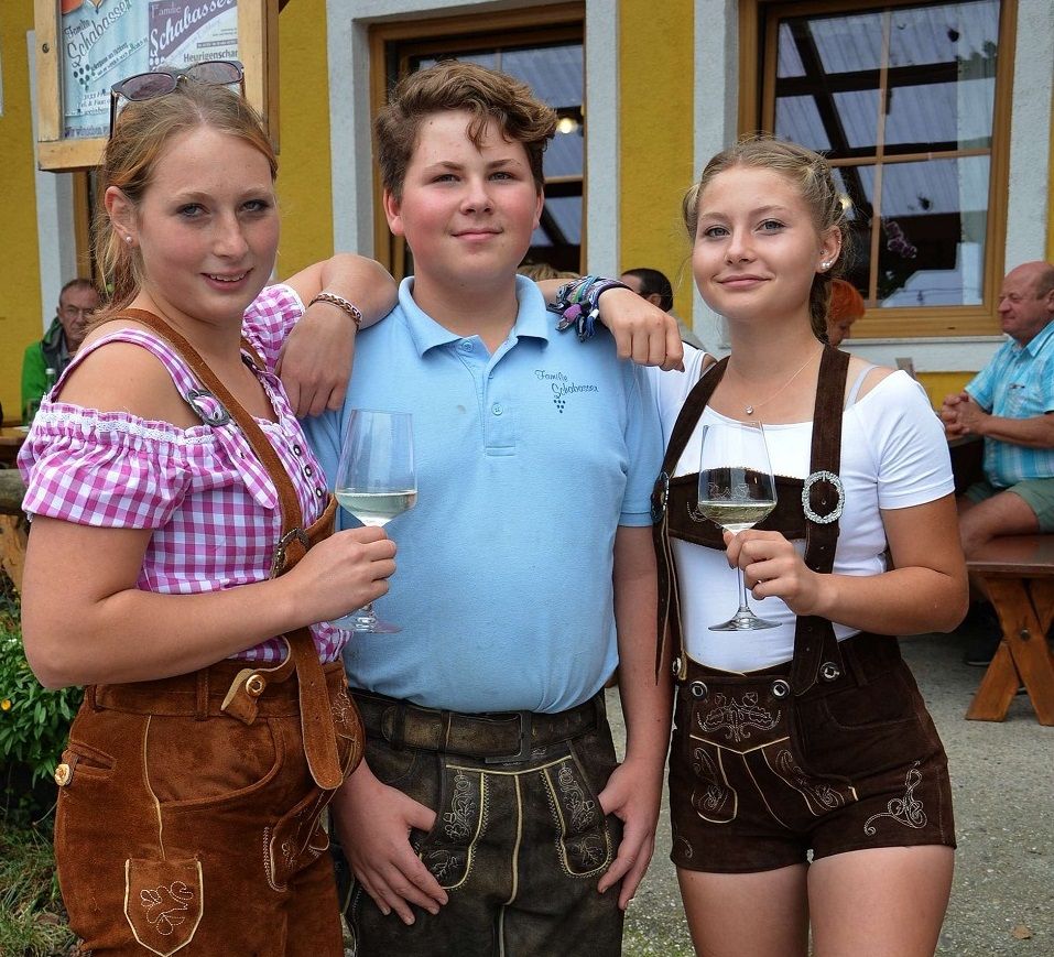 Three young people in traditional Bavarian dress with glasses of wine in front of a yellow building.