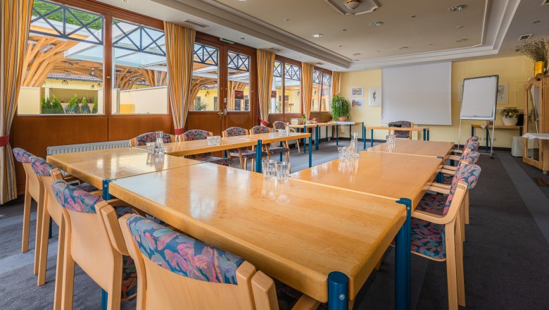 A bright seminar room with large windows, U-shaped tables, chairs and a flipchart.