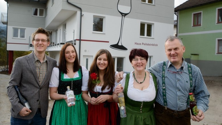 Krenn family, © Wirtshausbrennerei Krenn, Johann Frank A family stands in front of a building labeled 'Haus Peilsteinblick'. They are wearing traditional clothing and holding bottles in their hands.