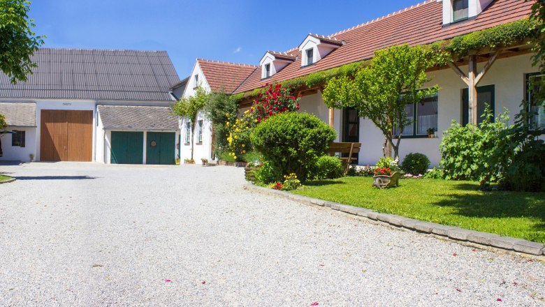 An idyllic vineyard with a gravel path, flowering gardens and traditional buildings under a blue sky.