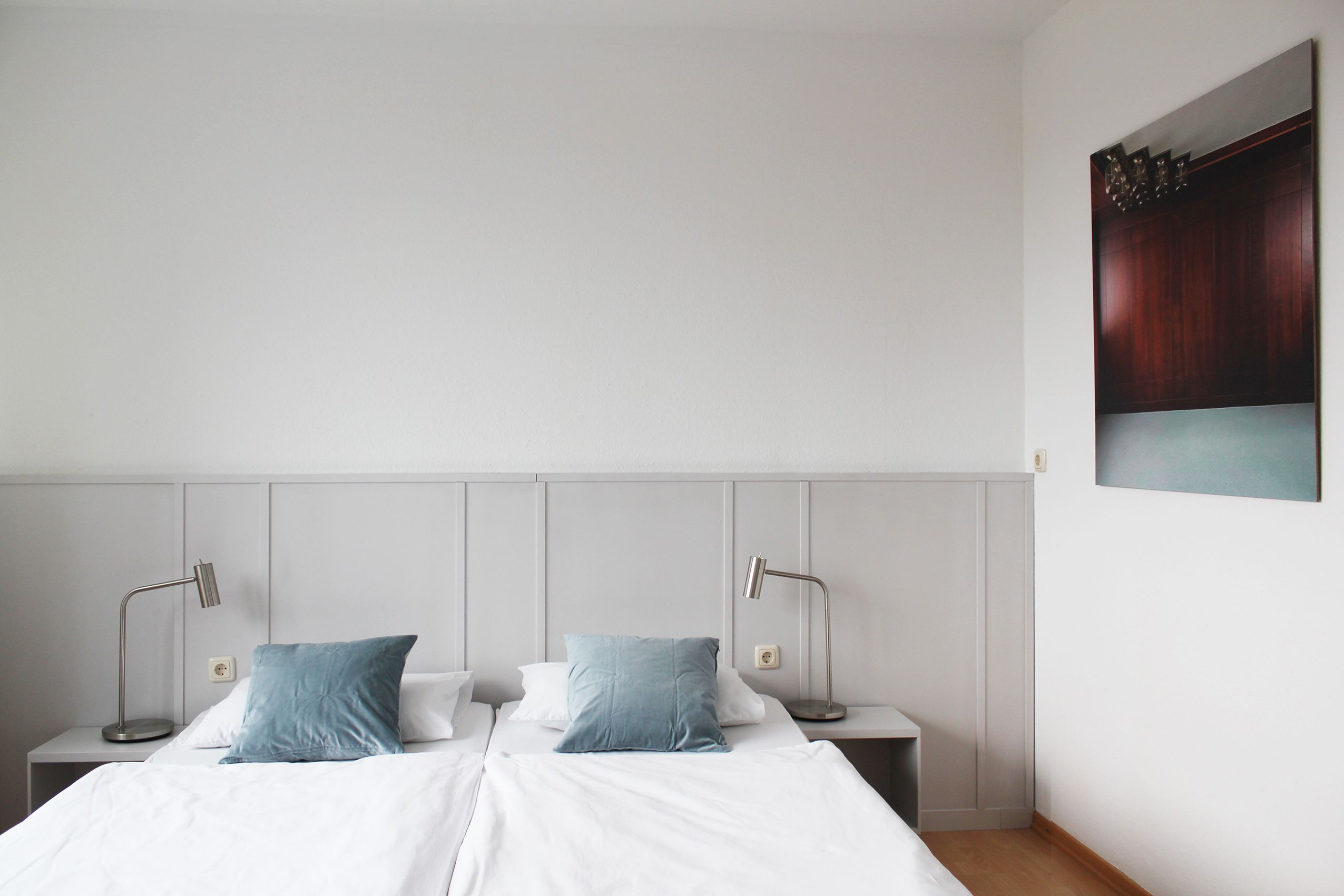 A simple guest room with a double bed, blue cushions, white walls and a picture on the wall.