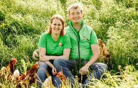 A man and a woman in green T-shirts sit smiling in a meadow surrounded by chickens.
