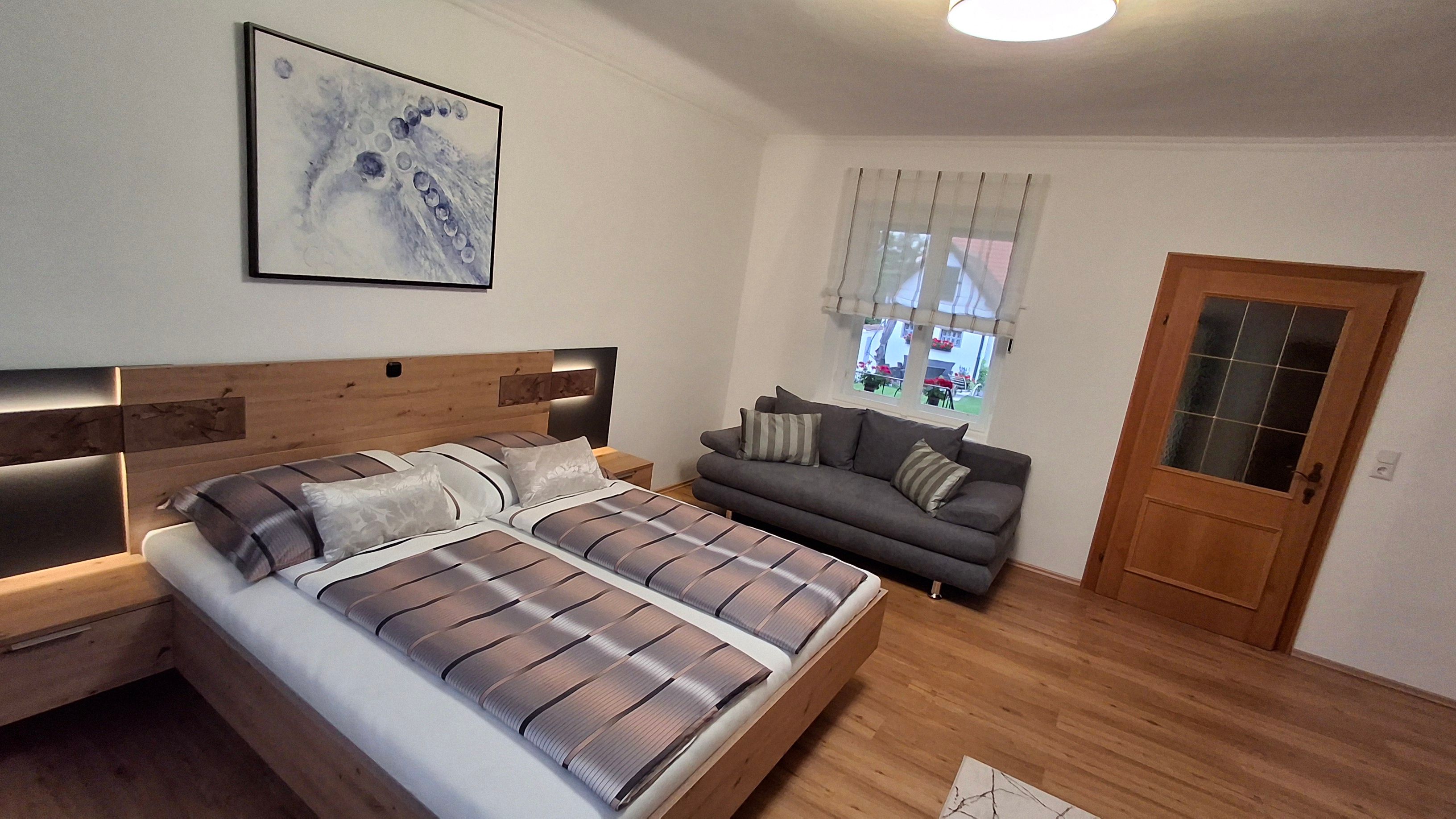 Modern bedroom with double bed, gray sofa, wooden floor and abstract artwork on the wall.