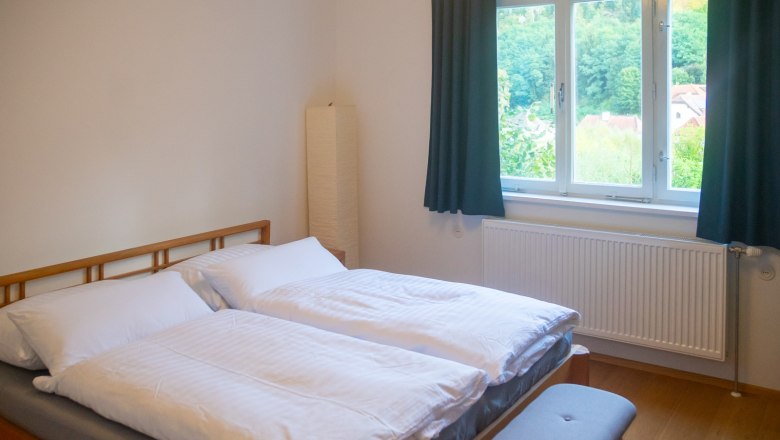 A cozy bedroom with a double bed, white comforters, a window with a view of the greenery and a radiator underneath.