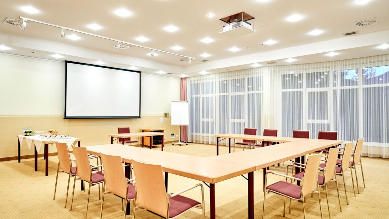 A bright seminar room with a U-shaped table arrangement, screen, flipchart and drinks table.