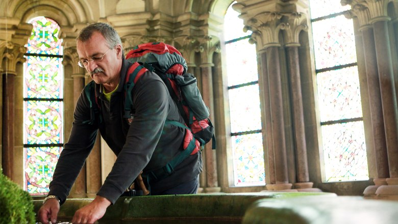 A man with a rucksack bends over a pool of water in a historic building with stained glass windows.