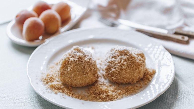 Two apricot dumplings on a plate with breadcrumbs, apricots in the background.
