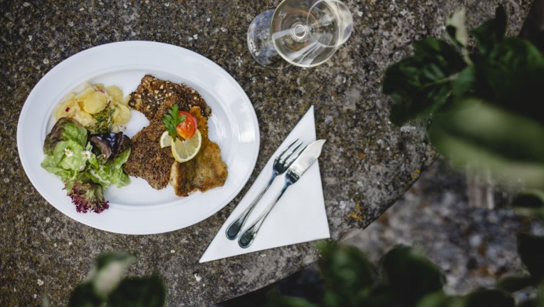 A plate with breaded fish, salad and potatoes on a stone table, cutlery and a wine glass next to it.